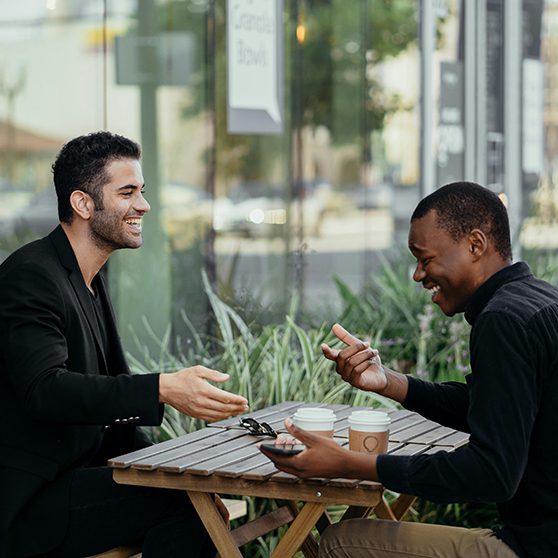 Two friends drinking coffee and laughing together at outdoor table.