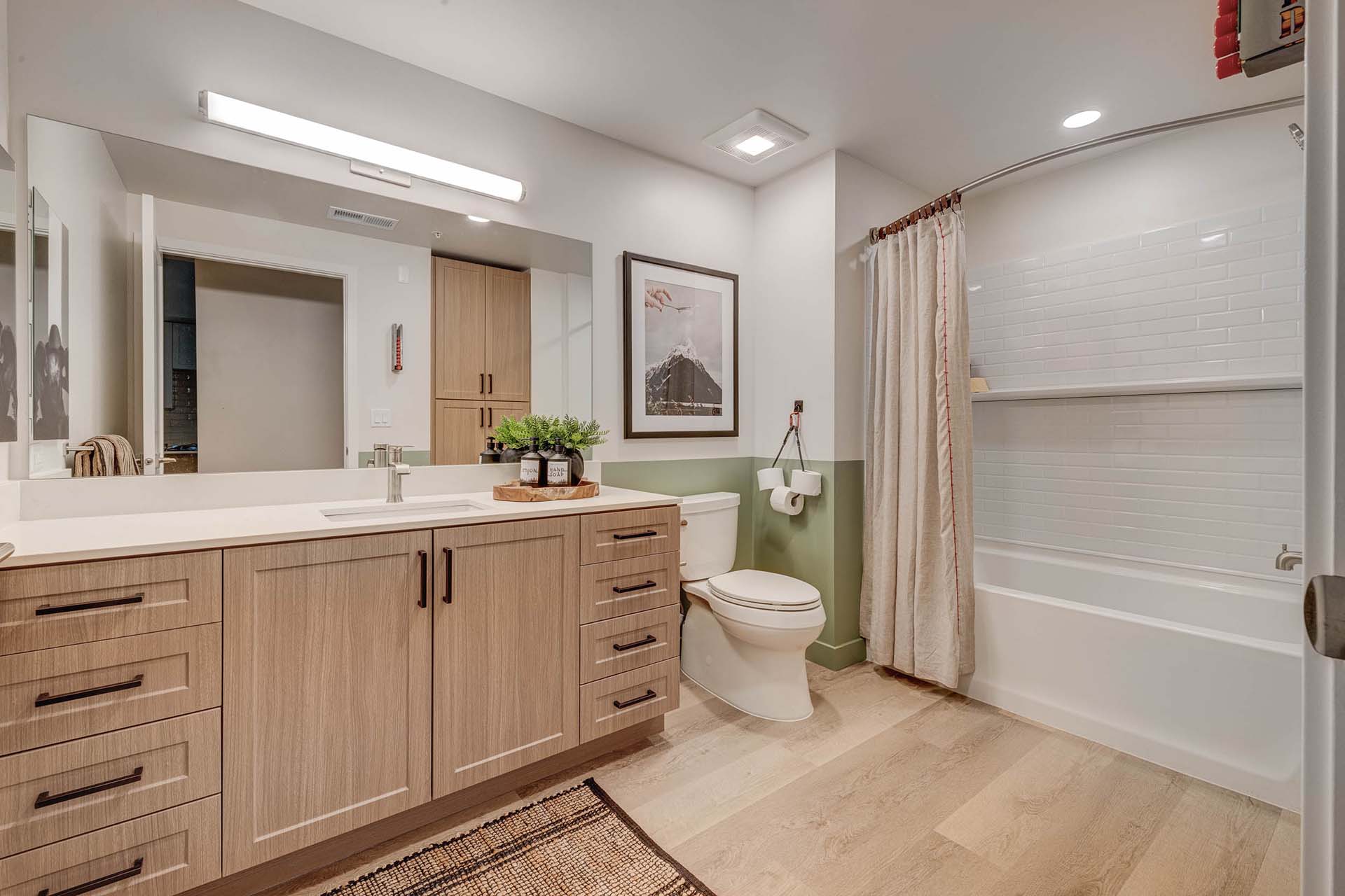 Millhaus apartment bathroom with wood style floor and cabinets, large mirror, and tiled shower tub.